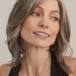 Woman with shoulder-length gray-blonde wig, natural makeup, smiling softly.